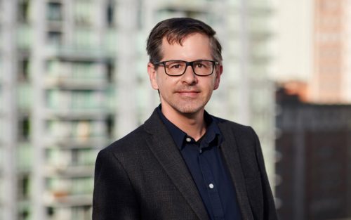 man with glasses smiling at the camera with a tall building in the background