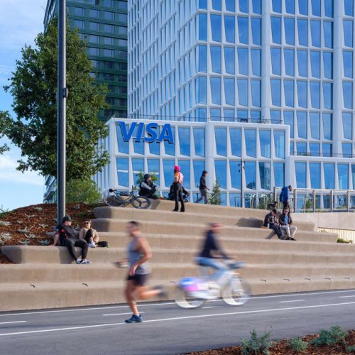 Visa office building closeup from the street with people sitting in front of the building, a man running and person cycling