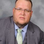 headshot of a man with a grey suit, blue shirt, and a green tie