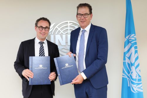 two spectacled men standing in front of a grey wall holding blue books, with the UN flag beside one the them