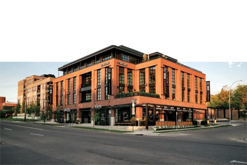 A contemporary brick building featuring several storefronts and a greenery line along the sidewalk. Streetlights stand beside the entrance, and the setting sun casts a warm glow over the scene.