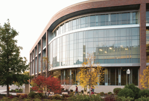 A contemporary building featuring large glass walls, curved edges, and several trees in various colors planted around it. Two people are seen walking along the path in the foreground, while the setting sun casts a warm light on the structure, enhancing its modern design.