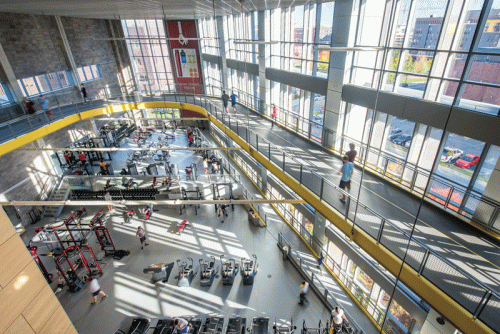 An expansive gym interior with large windows allowing natural light. The facility features workout areas with various equipment, such as weight machines and treadmills. People are exercising, some walking on a upper track that curves around the space.