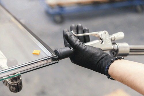 Person wearing black gloves using a sealant applicator tool to apply sealant along the edge of a glass panel in a manufacturing or assembly process. An orange spacer is visible on the glass panel.