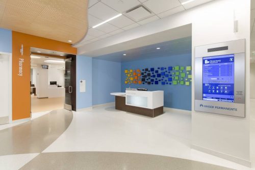 A modern reception area at Kaiser Permanente features a welcome desk, an orange sign indicating the pharmacy, and a digital directory on the right. The walls are painted in soft blue tones, and a colorful abstract display made of squares adds visual interest.