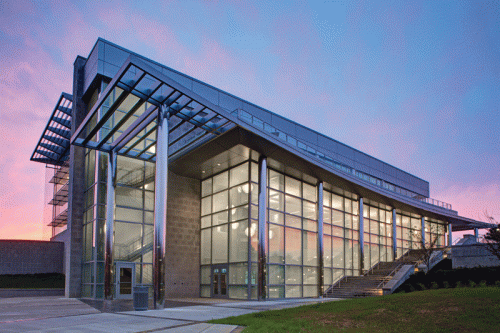 A contemporary architectural building featuring extensive glass walls and a distinctive angular roof. The entrance is highlighted by wide stairs leading up to the main doors. The backdrop displays a vibrant sunset with soft pink and purple hues.