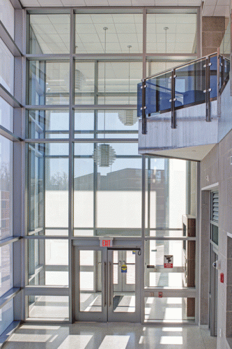 A bright, modern lobby features large glass windows allowing natural light. Double glass doors are centered at the bottom, with an "EXIT" sign above them. A balcony with glass railings is visible on the upper left, and light fixtures hang from the ceiling.