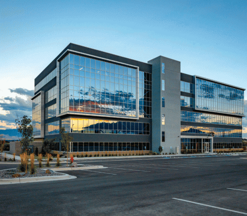 A contemporary office building features a sleek glass facade that reflects vibrant colors from the sunset and surrounding mountains. The parking lot is empty, with a few trees and shrubs in front of the building, enhancing its modern aesthetic.