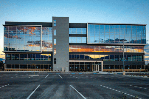 A contemporary office building with multiple floors features expansive glass windows reflecting a vibrant sunset sky and nearby mountains. The parking lot in front is empty, with clear lines marking designated spaces.