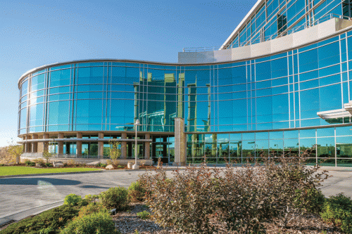 A contemporary, curved glass building features large windows reflecting the sky. It is set in a landscaped area with greenery and shrubbery in the foreground, showcasing a well-maintained outdoor space.