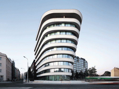 A contemporary architectural design featuring a curving white facade with multiple large windows. The building stands at a street intersection, surrounded by other structures, showcasing a modern urban environment.