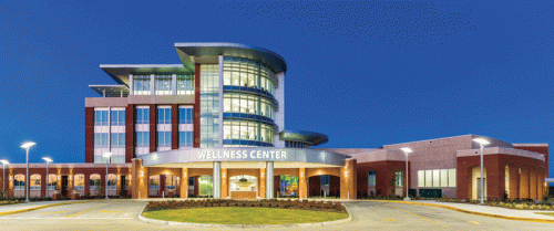 A contemporary wellness center featuring a large, multi-story structure with extensive glass panels, a prominent entrance sign reading "WELLNESS CENTER," and landscaping with illuminated pathways. The building is well-lit at twilight, showcasing its architectural features.