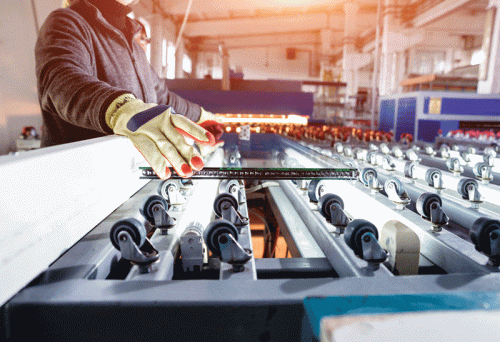 A factory setting with a worker handling glass panes on a roller conveyor system. The worker is wearing gloves for safety, and the machinery is illuminated under industrial lighting. The equipment includes multiple rollers designed for transporting glass panes.