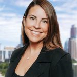 Professional headshot of Kristina Abrams with long brown hair, smiling, wearing a black blazer, with a cityscape and blue sky in the background.