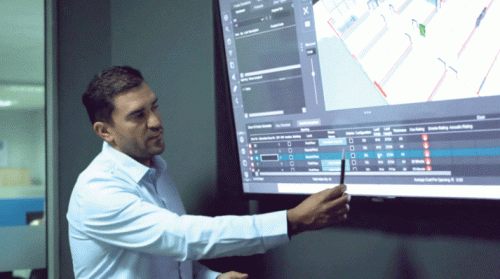 A man wearing a light blue shirt stands beside a large digital screen, pointing with a pen. The screen displays a 3D layout of a space, indicating various items with color-coded markers. Beneath the layout, there are several data tables with columns likely showing metrics and statistics, suggesting a technical presentation or analysis in a professional environment.