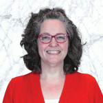 A close-up portrait of Amy Becker smiling in front of a marble-like background. She has curly gray hair, wears red-framed glasses, and a red sweater. Her bright smile conveys a friendly and approachable demeanor.