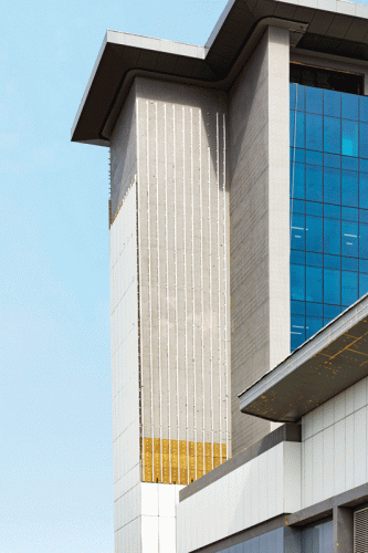 A contemporary multi-story building with a combination of smooth concrete and large glass windows. The structure displays a unique design with gold accents at the lower section. The top features a prominent overhanging roof, and the background is a clear blue sky.