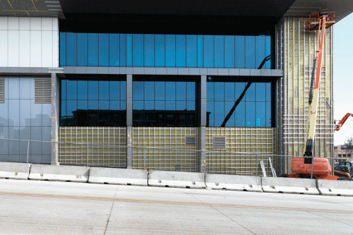 A construction site showcasing a modern building with large reflective glass windows. Scaffolding is visible on the right side, where a lift is positioned for work. Safety barriers line the bottom of the building, and the ground surface appears paved.