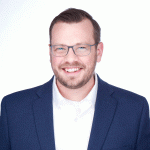 A smiling Benjamin Meyer with short hair and glasses stands against a bright white background. He is dressed in a navy blazer over a white collared shirt, conveying a friendly and professional demeanor.