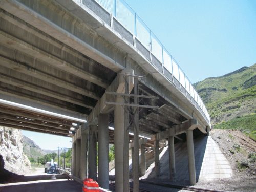 The image shows a concrete overpass viewed from below, supported by sturdy pillars. In the foreground, there's a red construction cone and a glimpse of a truck by the roadside. The backdrop features rolling green hills under a clear blue sky.