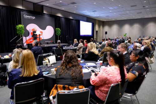 A professional conference setting featuring a speaker on stage addressing an audience seated at round tables. The stage has a modern design with the word "Coverings" displayed prominently, flanked by decorative plants. Attendees are engaged, with notepads, coffee cups, and other materials visible on the tables in a well-lit conference room.