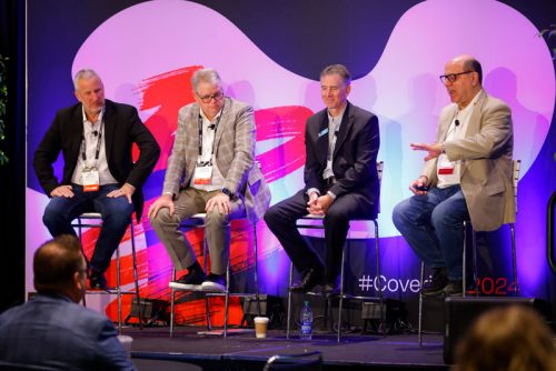 A panel discussion at a conference featuring four male speakers seated on high stools in front of a colorful, modern backdrop. The backdrop includes abstract designs and the word "Coverings." One panelist gestures while speaking, engaging both the audience and the other panelists. Water bottles, coffee cups, and conference badges are visible, indicating a formal yet conversational event.