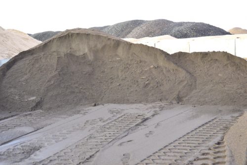 A large mound of gray sand, with distinct tire tracks visible in the foreground. In the background, there are rolling hills of varying sizes, creating a textured landscape under a clear sky.