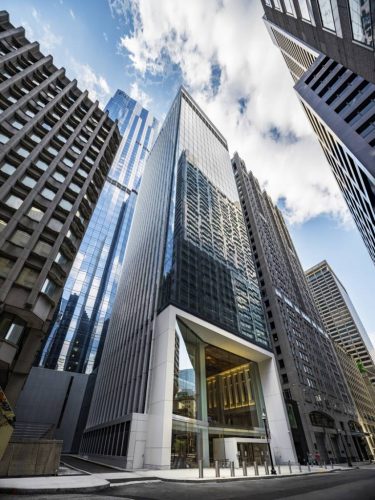 The image shows a modern glass skyscraper nestled between taller buildings. The structure features a large, transparent entrance and is surrounded by other architectural styles. The sky is partly cloudy, creating a dynamic atmosphere.