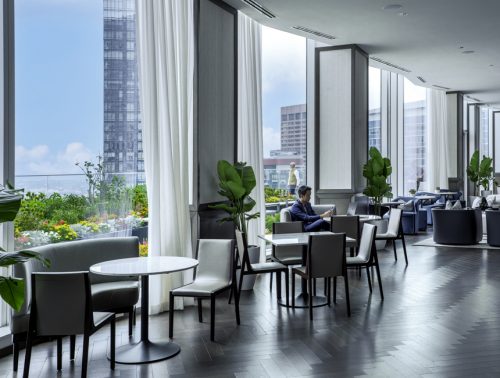 A sunlit lounge featuring large floor-to-ceiling windows, plants, and stylish furniture. A man sits at a table reading, while views of a cityscape and colorful flowers can be seen outside.