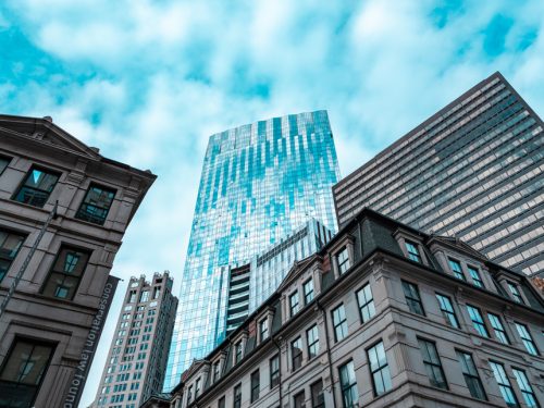 City skyline from a low angle, featuring a modern glass skyscraper on the right, reflecting clouds and sunlight. In contrast, an older, ornate building with a detailed facade is shown in the foreground on the left, creating a juxtaposition between contemporary and historical architecture.