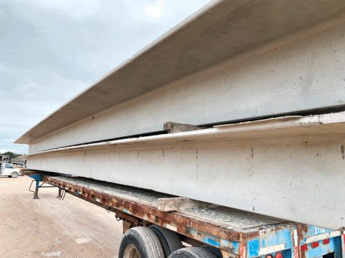 A trailer carrying several long concrete beams is parked on a dirt surface. The beams are arranged horizontally and appear heavy and sturdy. The scene is set under a cloudy sky, and there are hints of vehicles in the background.