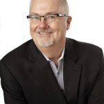 Professional headshot of Bill DuBois with short white hair, glasses, and a neatly trimmed beard, wearing a dark suit jacket and light-colored shirt, set against a plain white background.