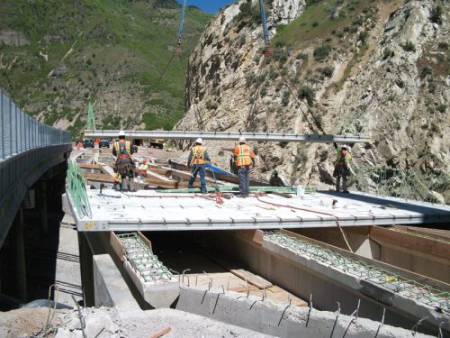 A group of construction workers in safety vests and helmets is positioned on a bridge being constructed over a deep valley. They are working on a concrete deck, with several workers engaged in various tasks, while cranes are used to lift materials. The rocky cliffs and green mountains are visible in the background, indicating a natural environment.