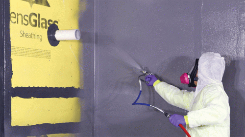 A worker wearing a white protective suit and mask is using a spray gun to apply a gray coating to a wall covered with yellow sheathing labeled "SensGlass." A white pipe is visible in the wall, and the environment appears industrial.