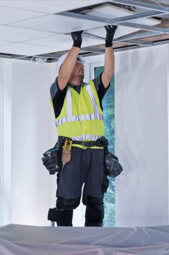 A worker in a bright yellow safety vest and helmet is installing ceiling panels in a spacious, well-lit room. The worker is using both hands to position the panel and is equipped with gloves. Tools hang from a belt around the waist. The room features large windows that allow natural light and has minimal furnishings.