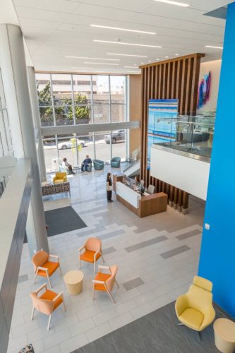 A spacious, modern lobby features a reception desk attended by a staff member. Bright orange and light blue chairs are arranged for seating. Large windows allow natural light and provide views of the street outside, where people can be seen sitting and cars are parked. The decor includes wooden panels and contemporary artwork, creating a welcoming atmosphere.