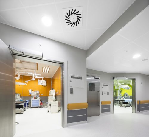 A well-lit hospital corridor featuring open doors on either side that lead to operating rooms. The walls are painted in neutral tones, with an orange accent wall visible through one door. Medical equipment can be seen in both rooms, suggesting a sterile and functional environment.