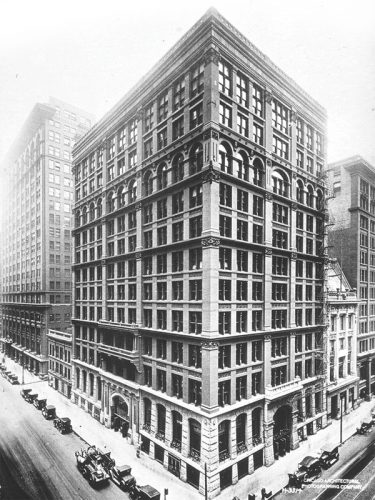 A historic black-and-white photograph showing a large multi-story building with ornate architectural details on its façade. The building is located at a city intersection, surrounded by other tall structures, with early 20th-century automobiles parked along the street below. The image captures the bustling atmosphere of the urban environment during that era.