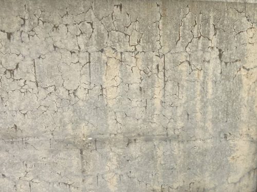 A close-up view of a concrete wall displaying a pattern of cracks and textures. The surface is mostly gray with lighter areas, giving it an aged appearance.