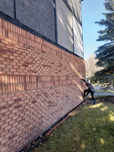 A person in a gray sweater is climbing a brick wall of a building, positioned on the left side of the image. The wall is made of reddish-brown bricks and is surrounded by a green lawn and trees. In the background, there is a clear blue sky.