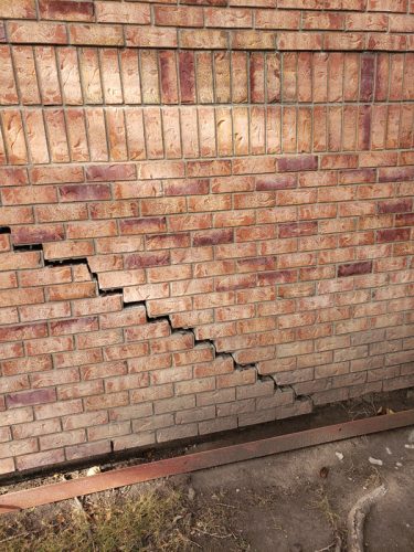 A brick wall displays a significant diagonal crack running from the bottom left to the upper right. The bricks are various shades of red and brown, with some mortar visible between them. The crack disrupts the pattern of the bricks, creating an uneven appearance. Below the wall, a metal structure and dirt can be seen, indicating potential issues with the foundation.