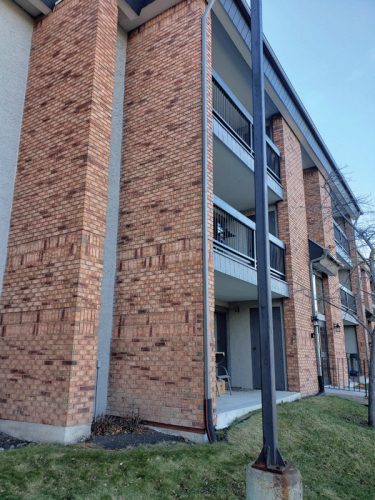 A corner view of a three-story apartment building featuring a brick exterior. The building has several balconies with black railings on the second and third floors. Below the balconies, there is a small area of grass and a support pole in the foreground.