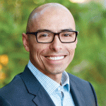 Larry Fosnight with a shaved head and glasses smiles warmly at the camera. He is wearing a dark suit over a light blue checkered shirt. The background is softly blurred, featuring greenery, indicating an outdoor setting.