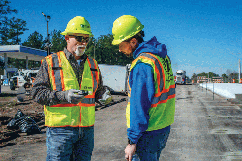 Two construction workers are engaged in conversation on a work site. One, wearing a patterned shirt under a safety vest, holds a measuring device, while the other, clad in a blue hoodie and safety vest, looks down at it. In the background, construction equipment and a clear blue sky are visible.