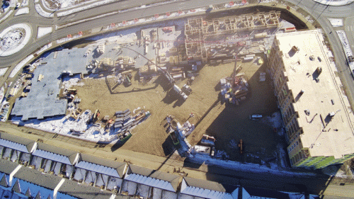 An aerial perspective of a large construction site featuring various heavy machinery. The site includes a large area of bare ground, stacks of materials, and partially constructed buildings. To the right, a building is in the final stages of construction, while the left side shows an area with a solid foundation and machinery in use. Surrounding roads and some snow-covered areas are visible.