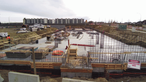 A construction site with a foundation poured and framed. Workers are moving materials around while construction vehicles are visible. In the background, apartment buildings stand. A "Construction Area" sign warns of work in progress.