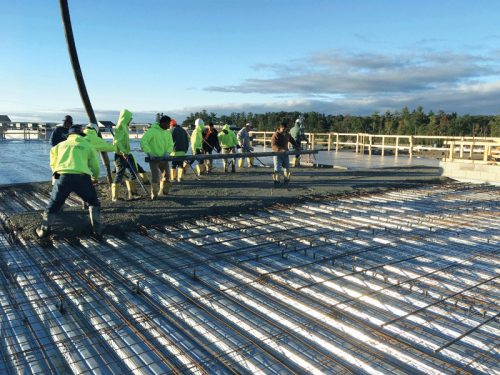 A group of construction workers wearing bright yellow rain jackets and boots is pouring concrete onto a rebar framework. The workers are actively engaged in the process, with one person using a long screed to level the concrete. The work occurs on a flat surface with wooden structures in the background, under a clear blue sky with a few clouds.