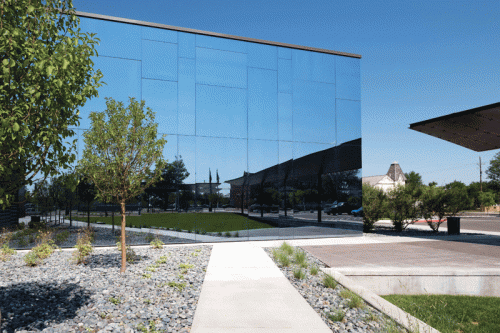 A contemporary glass building with a reflective surface stands prominently, surrounded by a landscaped area featuring trees and a stone pathway. The scene is set against a clear blue sky, with green grass visible in front of the building and a compact design incorporating shrubs and pebbles.