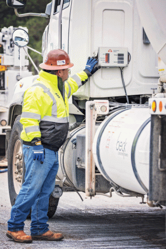 A construction worker wearing a hard hat and a reflective yellow jacket stands next to a truck, interacting with a control panel on its side.