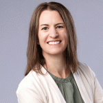 Lisa May with medium-length brown hair smiles warmly at the camera. She wears a light beige cardigan over a green blouse. The background is a soft gray, creating a professional and inviting atmosphere.